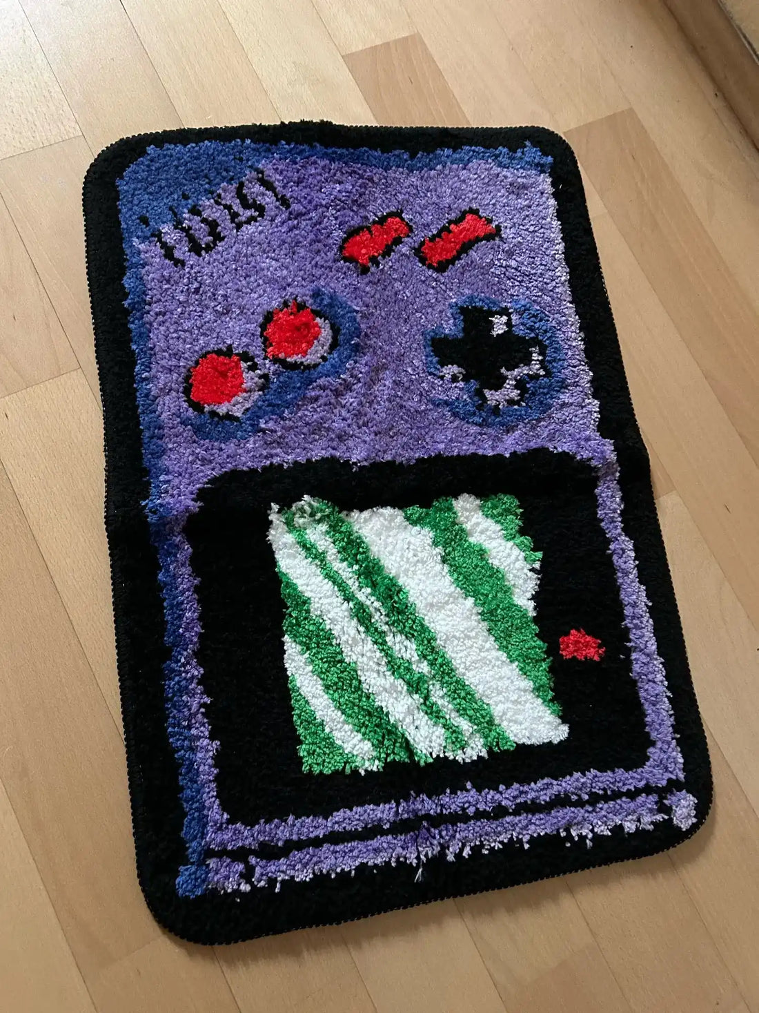 Tapis fait main en forme de Game Boy violet avec un écran à rayures vertes et blanches.