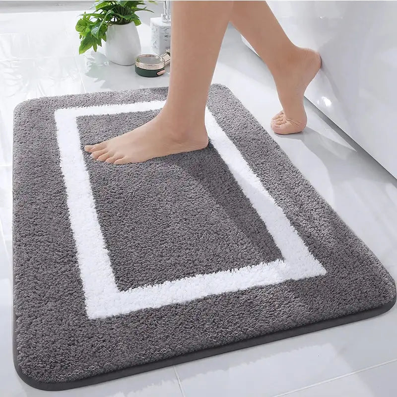Tapis de bain gris avec bordure blanche et surface texturée, probablement fabriqué en microfibre ou matériau similaire.