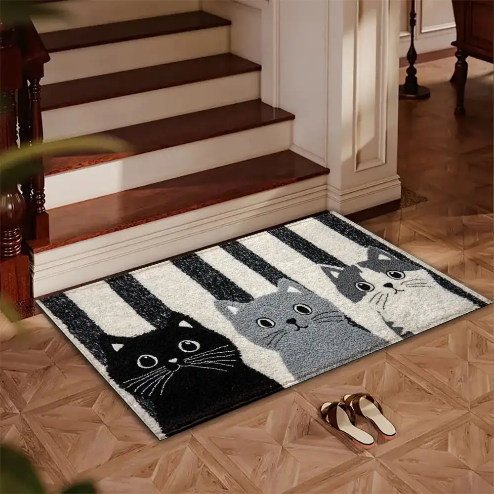 Un paillasson d’entrée rectangulaire présentant trois chats stylisés en noir, gris et blanc sur un fond rayé.
