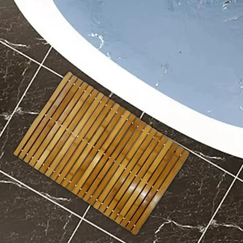 Un tapis de bain en bambou rectangulaire de couleur brun clair naturel et à conception en lattes, posé sur un sol carrelé sombre à côté d’une baignoire.