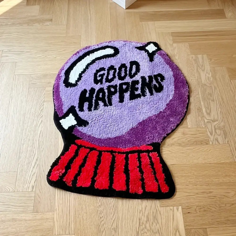 Tapis de bain boule à neige violette avec base rouge et texte blanc ’good happens’ sur sol en bois.