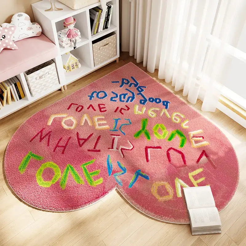 Tapis rose en forme de cœur avec lettrage coloré et design ludique, adapté aux chambres d’enfants ou aux aires de jeux.