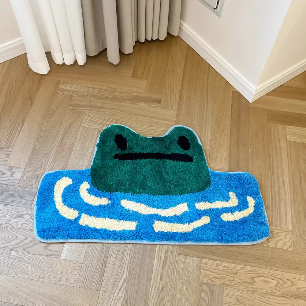 Un tapis de bain moelleux bleu orné d’une grenouille verte aux yeux noirs et à la bouche simple, sur un fond texturé de motifs ondulés blancs.
