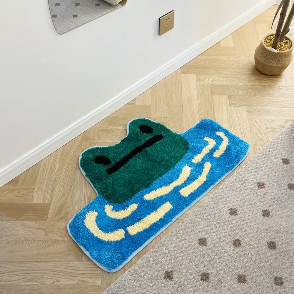 Un tapis de bain moelleux, style cartoon, en forme de grenouille verte avec des motifs de vagues bleues et beige.