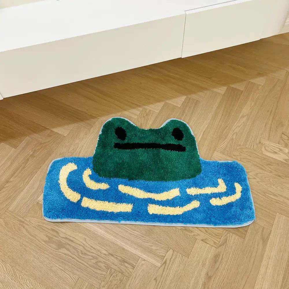 Un tapis de bain moelleux bleu orné d’une grenouille verte aux yeux noirs et à la bouche simple, entourée de lignes ondulées jaunes évoquant de l’eau.