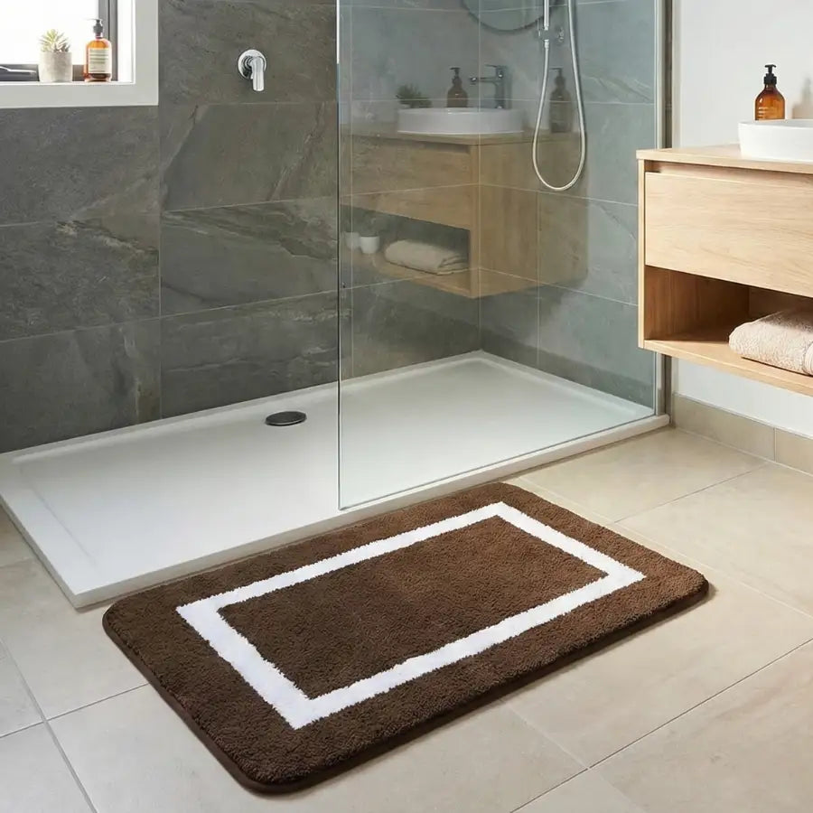 Tapis de bain rectangulaire marron avec bordure blanche et accents aux coins, probablement fabriqué en microfibre ou matériau doux similaire.