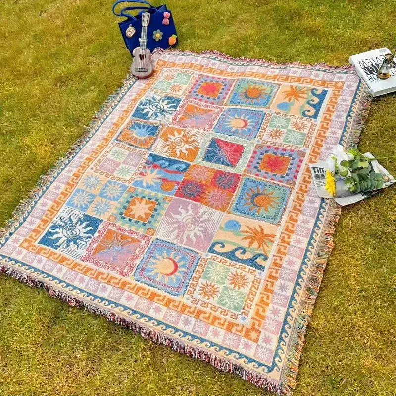 Une couverture en tapisserie tissée colorée présentant un design patchwork de soleils, lunes et palmiers en teintes vives comme l’orange, le bleu, le rose et le vert, avec des bords frangés.