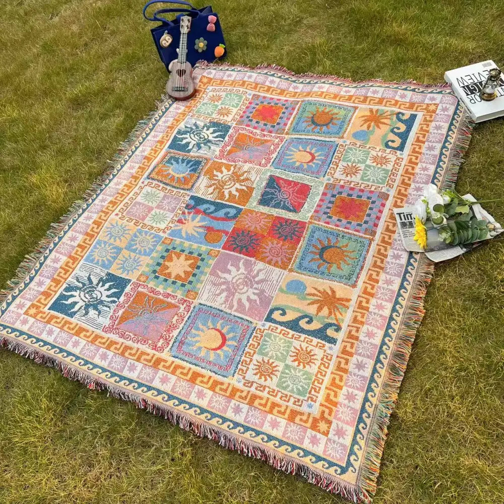 Une couverture en tapisserie tissée colorée présentant un design patchwork de soleils, lunes et palmiers dans des teintes vives comme l’orange, le bleu, le rose et le vert, avec des bords frangés.