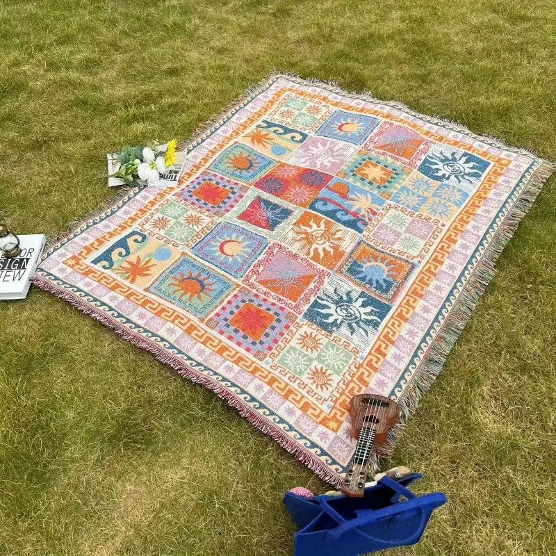 Une couverture tissée colorée en forme de tapisserie mettant en vedette un design patchwork de soleils, lunes et motifs géométriques dans des teintes vives comme l’orange, le bleu, le rose et le vert, avec des bords frangés.
