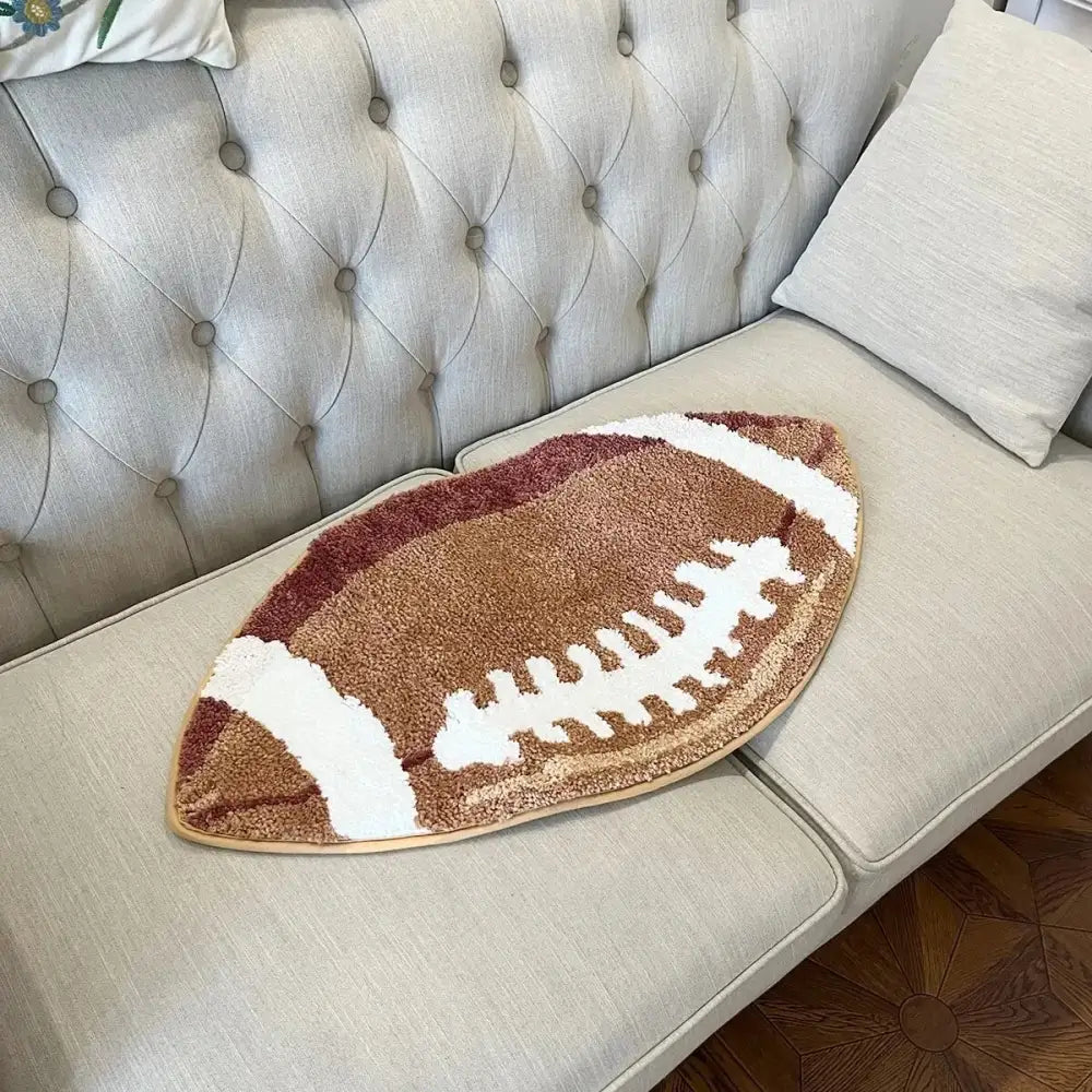Tapis brun en forme de ballon de football avec lacets blancs et bordure dorée, probablement d’une marque de décoration intérieure.