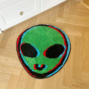 Tapis en forme d’alien vert avec des yeux noirs, une bouche souriante soulignée en rouge et bleu, bordé de cercles concentriques rouges, bleu clair et noirs.