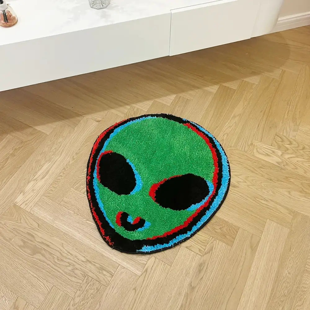 Un tapis de bain moelleux, circulaire, à thème d’alien vert avec des yeux noirs et une bouche souriante soulignée en bleu et rouge.