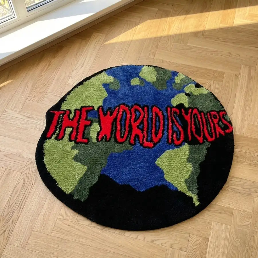 Tapis rond avec motif de carte du monde, continents verts et bleus sur fond noir, texte rouge « le monde est à vous »assistant : « tapis rond présentant une carte du monde vibrante en nuances de vert et de bleu sur un fond noir élégant, orné de