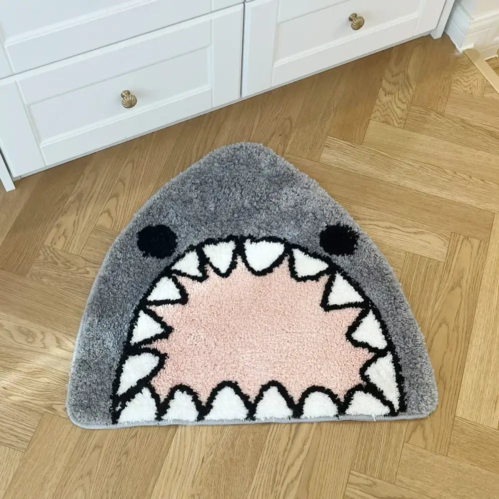 Tapis de bain gris en forme de requin avec des yeux noirs, des dents triangulaires blanches délimitées en noir, et une zone intérieure de bouche rose.