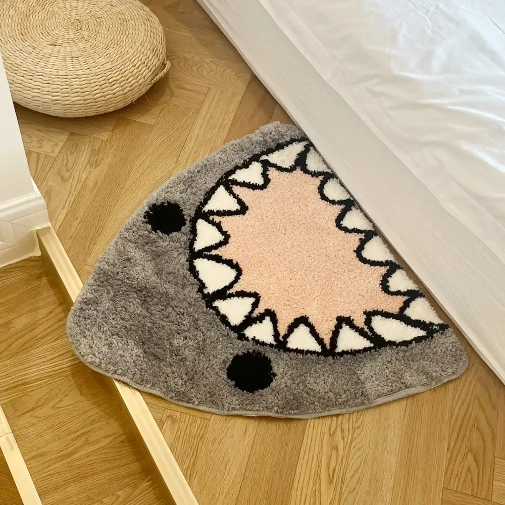 Tapis de bain triangulaire en peluche grise orné d’un design ludique de bouche de requin avec des dents blanches et des contours noirs, accentué par deux yeux circulaires noirs.