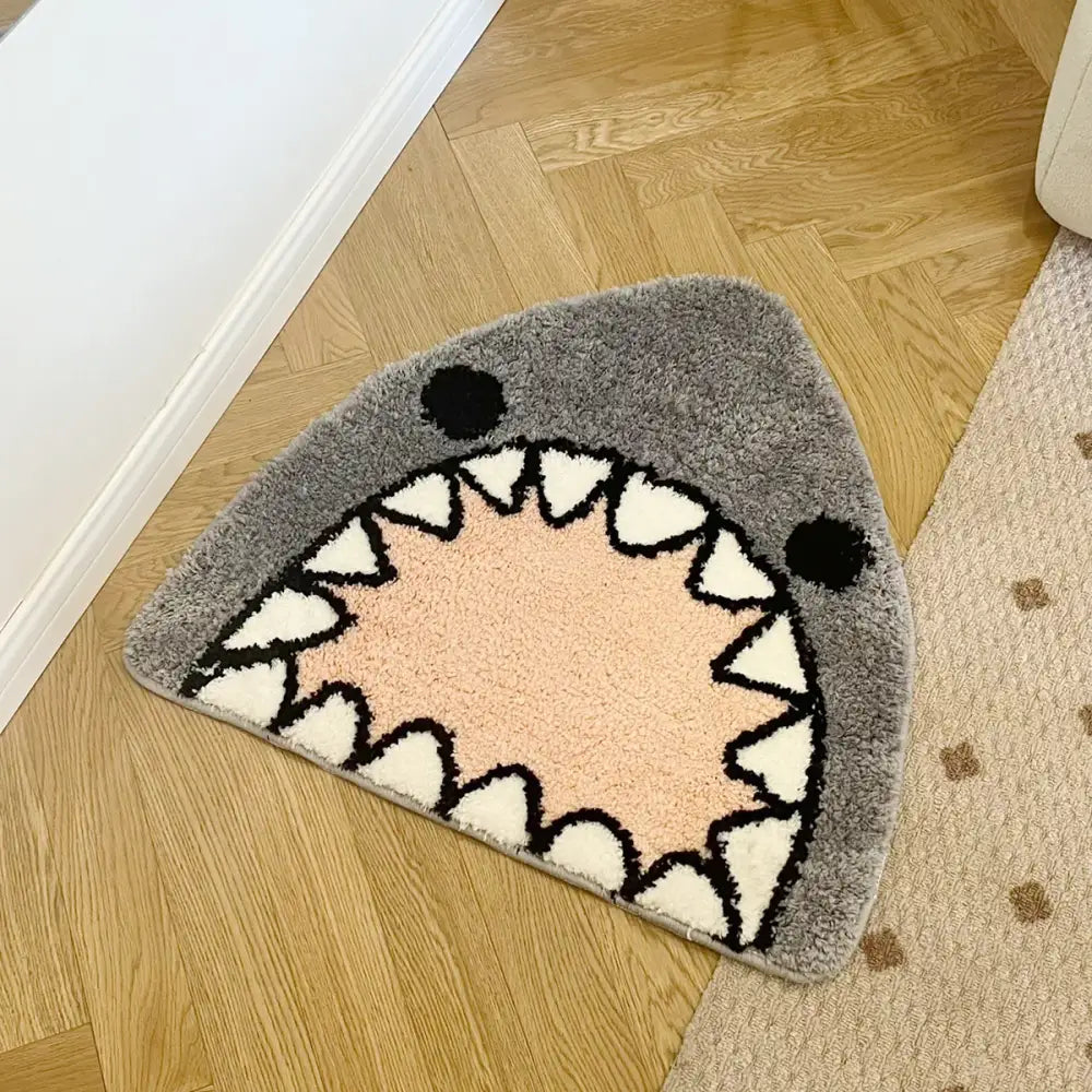 Tapis de bain en peluche grise en forme de requin avec des dents triangulaires blanches et des yeux circulaires noirs.