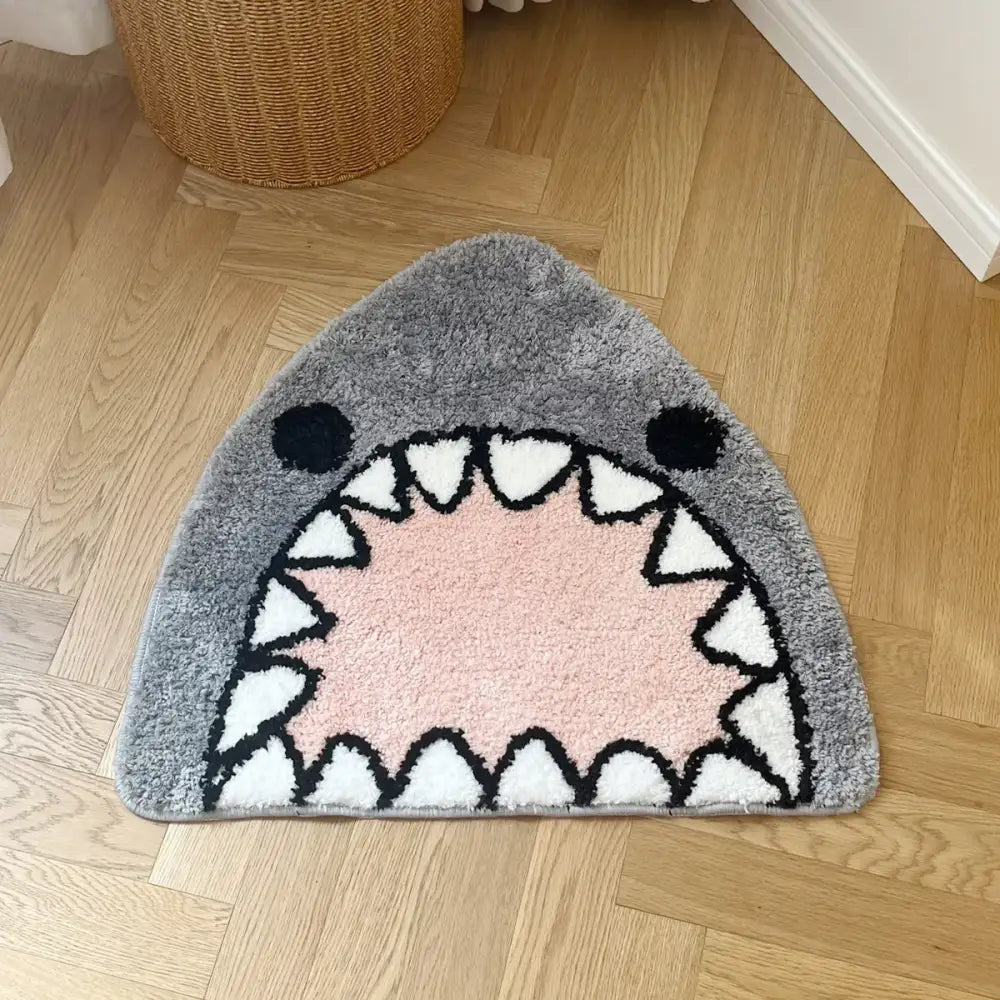 Tapis de bain gris en forme de requin avec un intérieur rose et des dents triangulaires blanches bordées de noir.