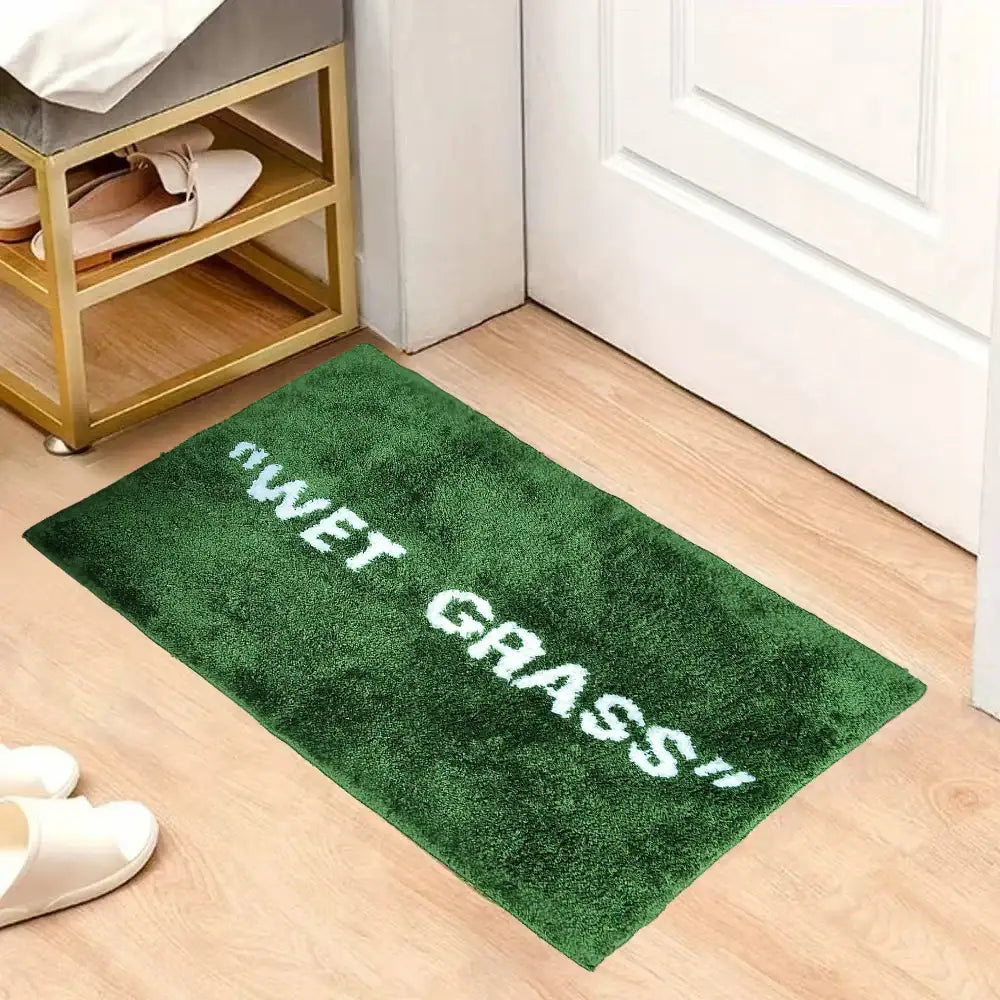 Paillasson en peluche verte avec du texte brodé en blanc indiquant « herbe mouillée ! »