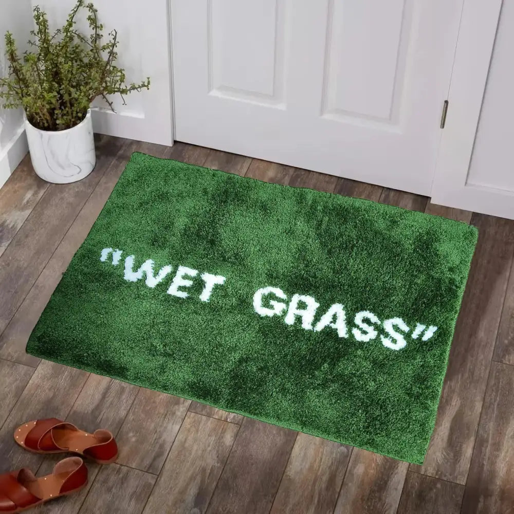 Paillasson en peluche verte avec du texte brodé blanc indiquant ’herbe mouillée’.