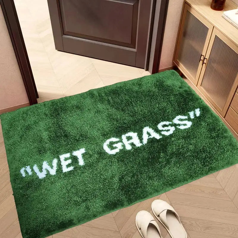 Paillasson en peluche verte avec du texte brodé blanc indiquant « herbe mouillée ».