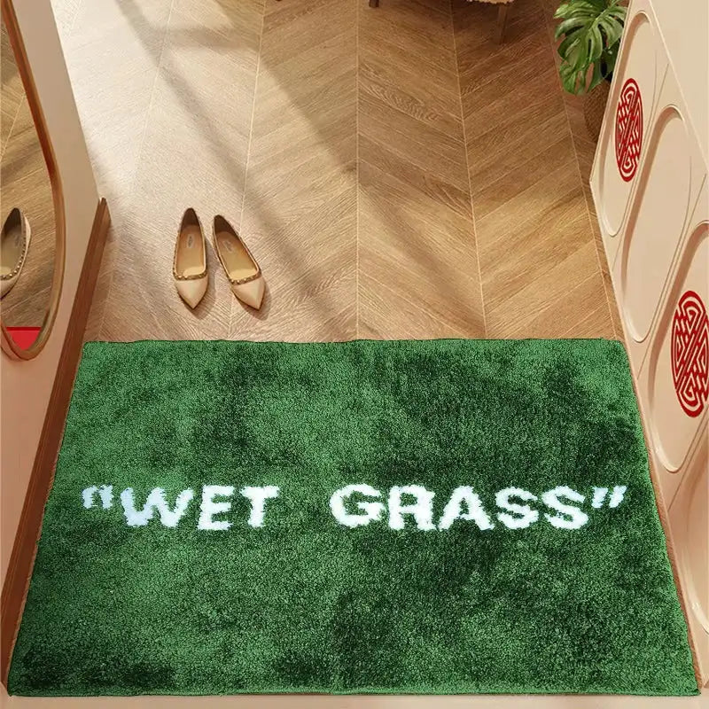 Paillasson en peluche verte avec ’herbe mouillée’ brodée en blanc.
